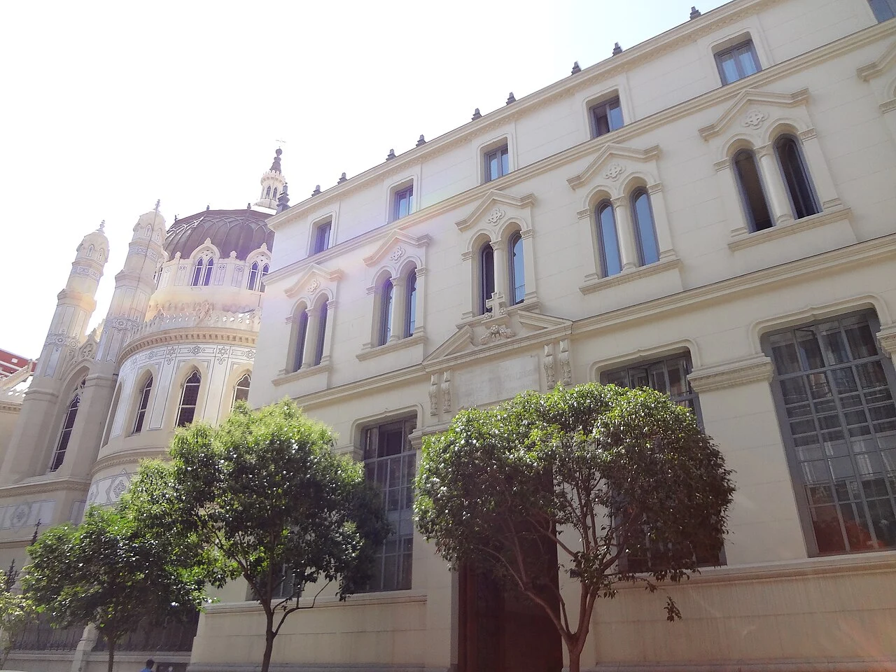 A pale, ornate historic building with arched windows and a decorative dome in Madrid, with two small green trees in the foreground.
